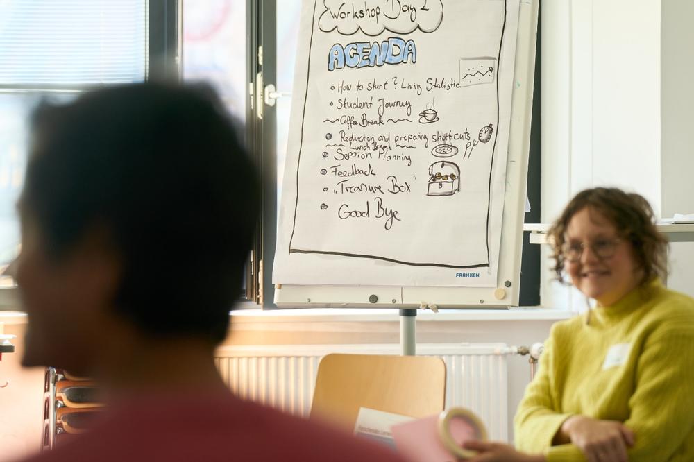 Workshopteilnehmende diskutieren im Vordergrund; im Hintergrund ein Flipchart mit der Agenda/Workshop participants engaged in discussion in the foreground; a flipchart with the agenda is visible in the background.